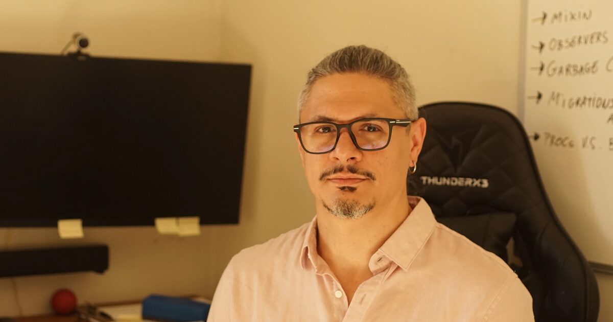 A man with short gray hair, glasses, a small goatee, and an earring, wearing a light pink button-up shirt, seated in an office chair with a monitor and whiteboard in the background.