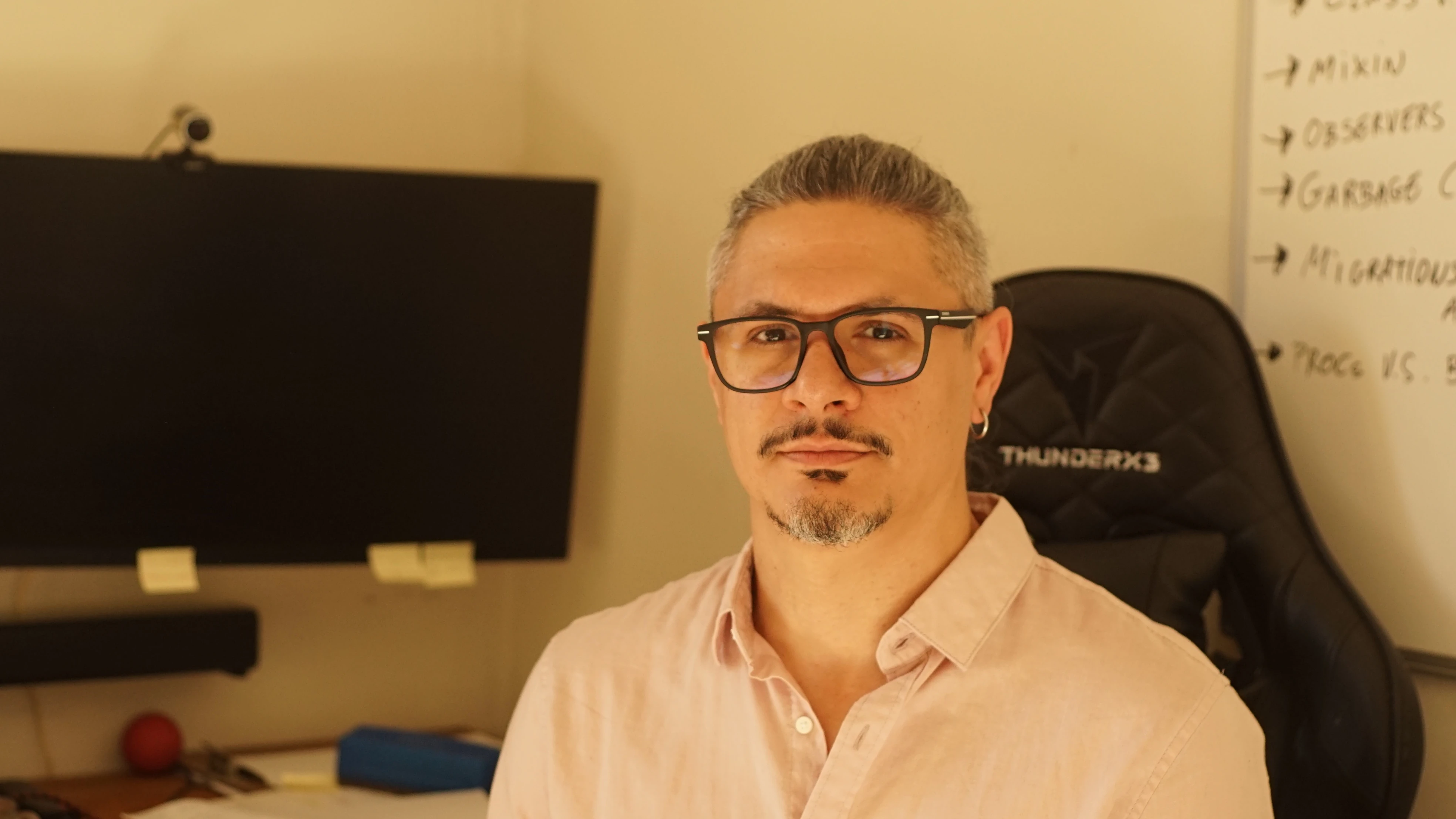 A man with short gray hair, glasses, a small goatee, and an earring, wearing a light pink button-up shirt, seated in an office chair with a monitor and whiteboard in the background.